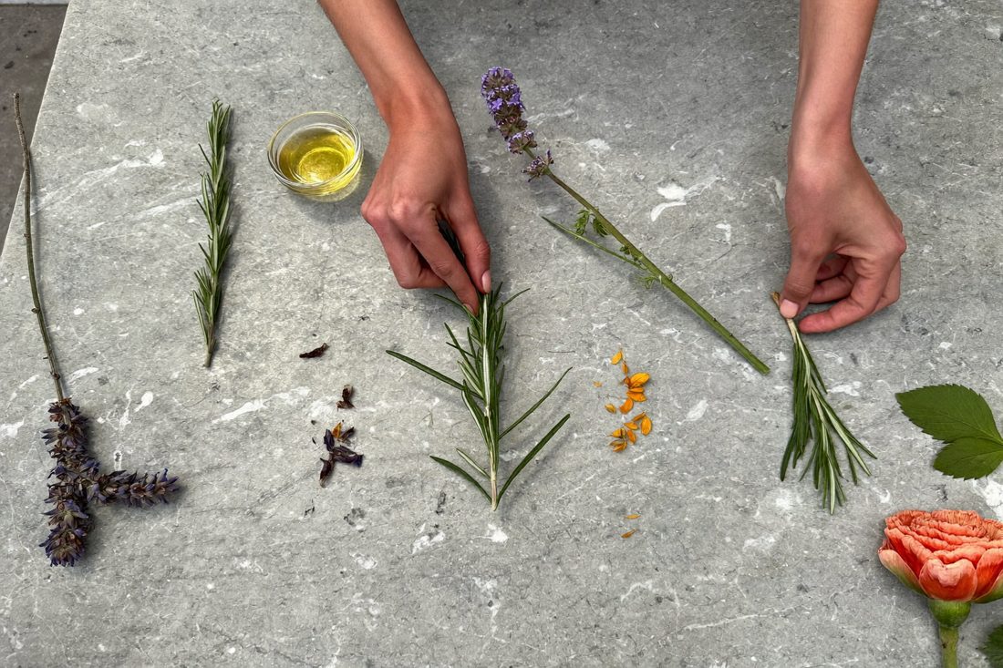 Mani che dispongono rametti di rosmarino, lavanda e fiori d'arancio su un piano di pietra
