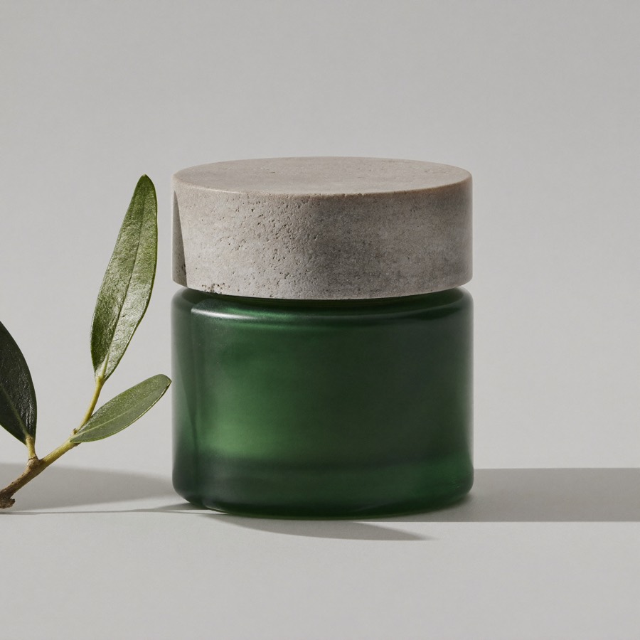 Vaso in vetro verde scuro con coperchio in pietra della Crema Ricca all'Ulivo accanto a un rametto di olivo